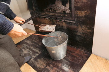 Senior man cleaning ash from a home fireplace with a metal shovel and bucket.