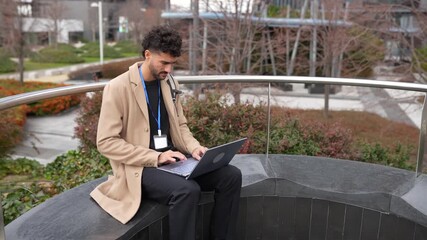 Youthful male doctor with a stethoscope is seated on a bench in a contemporary city park, diligently working on his laptop - Powered by Adobe