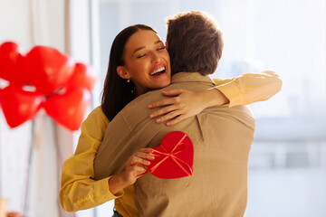 Happy woman smiling and hugging man while holding heart-shaped gift box, red balloons fill the room, couple celebrating anniversary or Valentine's day