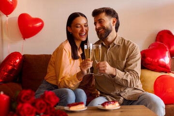 Happy man and woman raising their glasses in toast, having romantic dinner, celebrating anniversary or Valentines day, sitting on sofa with red balloons around