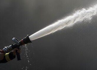 Firefighter's hand holding a fire hose nozzle, spraying a powerful jet of water. Dynamic water droplets and a strong stream against a dark background.