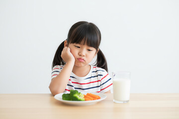 Unhappy little Asian girl refusing to eat healthy vegetables and milk, showing dislike for nutritious food.