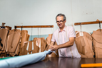 Professional male tailor working with light blue fabric in a sewing workshop with patterns hanging on the wall.