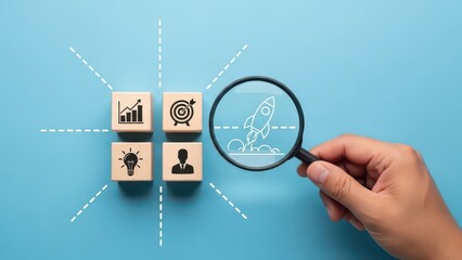 Hand holding magnifying glass over wooden blocks with business icons on a blue background