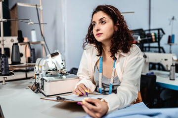 Young female fashion designer or seamstress sitting at an industrial sewing machine in a workshop, holding a clipboard and looking thoughtful while planning a new garment design.