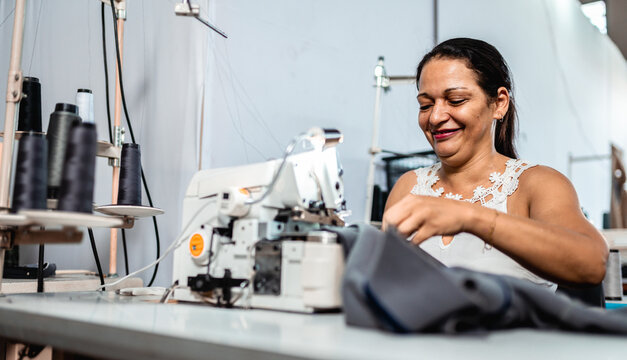 Smiling mature woman working as a seamstress in a garment factory, using an industrial sewing machine to stitch fabric in a bright workshop.