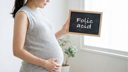 Pregnant woman holding a chalkboard with folic acid message, expecting mother in casual attire by a window, prenatal health and vitamin awareness in maternity care concept