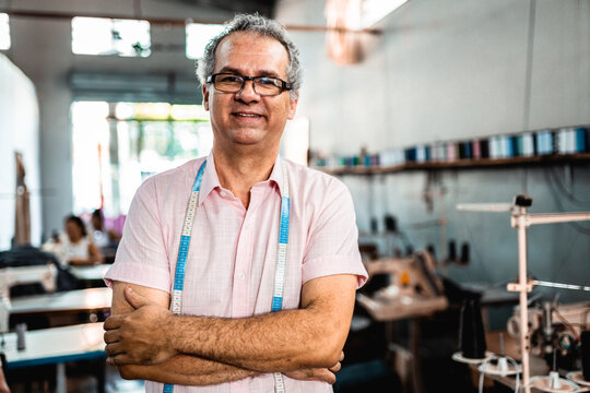 Portrait of a confident mature male tailor standing with arms crossed in a garment factory workshop, wearing a measuring tape and smiling at the camera.