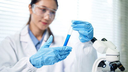 Asian adult woman scientist conducts laboratory research using microscope holding test tube blue liquid chemical analysis