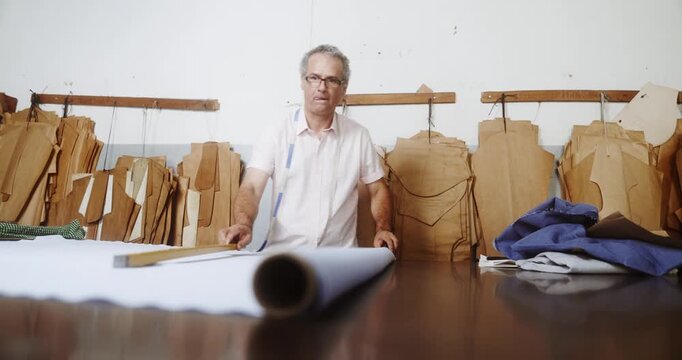 Professional mature tailor working with white fabric on a cutting table in a garment workshop with hanging patterns.