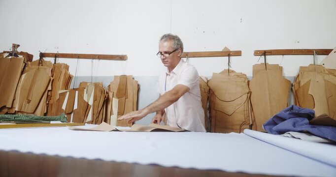 Professional mature tailor working with white fabric on a cutting table in a garment workshop with hanging patterns.