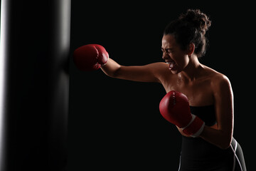 Angry female African-American boxer punching bag on black background