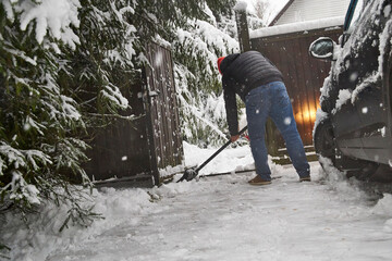 Man clearing snow near a car in winter yard, using a snow shovel during snowfall. Outdoor seasonal work, cold weather, driveway cleaning and winter home maintenance
