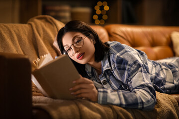 Obraz premium Beautiful young woman reading book and lying on sofa in living room at night