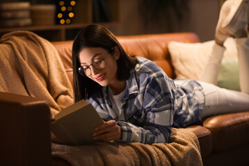 Obraz premium Beautiful young woman reading book and lying on sofa in living room at night