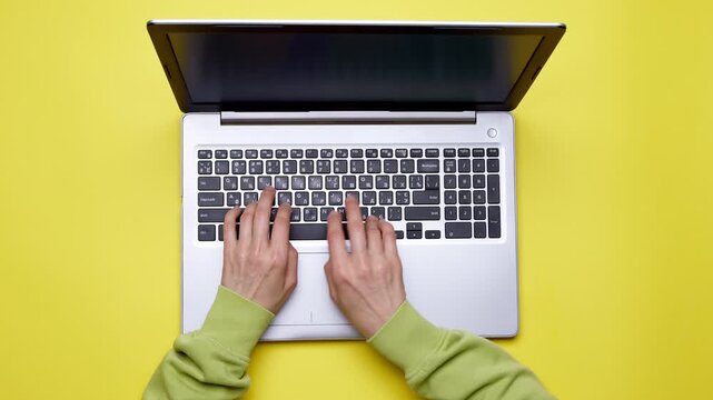 Top view. Hands typing on a laptop keyboard. Yellow background. Working on a computer at a desk.