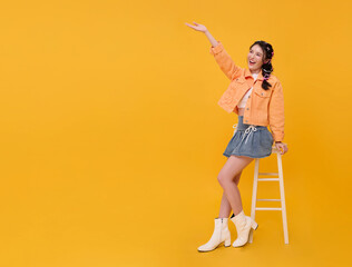 Lifestyle presentation promotion Asian teen woman sitting on chair and raising hand to showcase copy space cheerful gesture creative advertising modern youthful energy on yellow background.