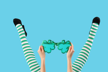 Female legs in green stockings with clover shaped novelty glasses on blue background. St. Patrick's...