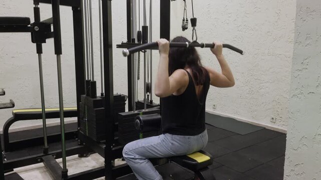 Back view of caucasian adult woman performing lat pulldown on gym machine indoors, arms raised and focused posture representing strength training and fitness routine.