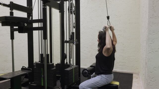 Caucasian adult woman sitting and performing lat pulldown exercise on gym machine indoors, focused posture and controlled movement symbolizing fitness training and healthy lifestyle.