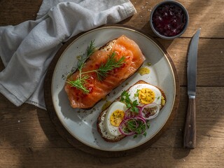 A rustic wooden table set with a white cloth, featuring a plate of smoked salmon, a poached egg with red onion and herbs, and a small bowl of jam. A knife and a spoon are placed beside the plate.