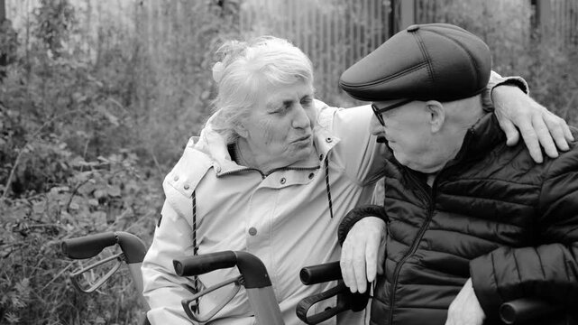 Elderly couple with grey hair and glasses sitting on walkers taking a break during walk in park. Hugging, laughing and having fun together