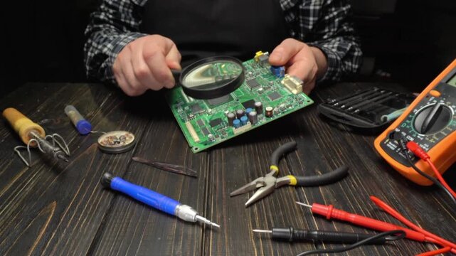 A person looks closely at a circuit board using a magnifying glass. Various tools like pliers and a multimeter are arranged around on a work table. This is a workspace for electronics repair.