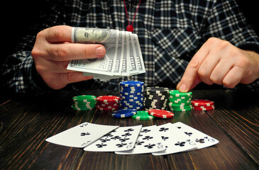 A person sits at a table with various colored poker chips and playing cards, holding cash in one hand. The focus is on the game preparation and betting
