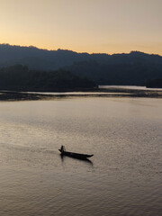 Silhouette of person rowing small wooden boat on calm lake water during golden sunset with dark mountain hills in background and orange sky reflecting on surface in peaceful evening atmosphere