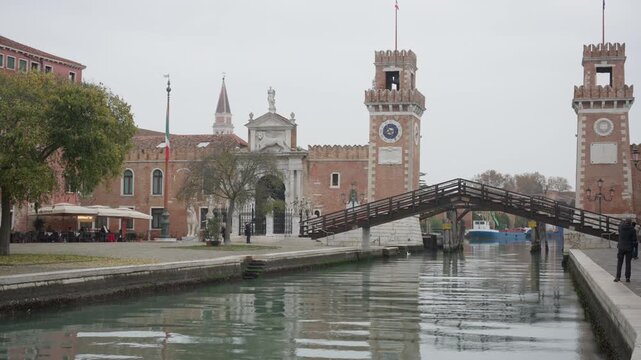 Venetian Arsenal entrance gate with historic brick towers and wooden bridge over the canal. Tourists walking in Venice Italy near Porta Magna under cloudy sky during day trip or vacation.