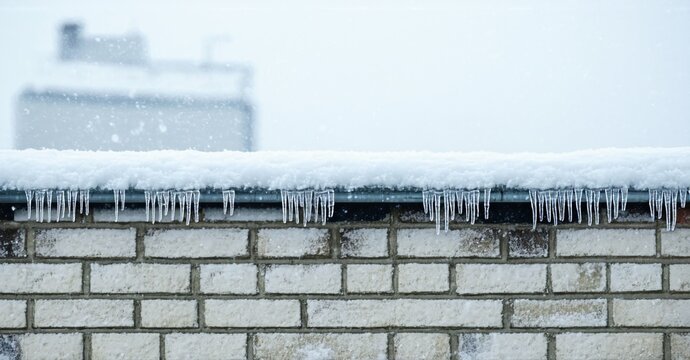 Winter scene shows icicles hanging from roof edge. Brick wall below, blurred buildings visible background. Snowy conditions present. - Powered by Adobe