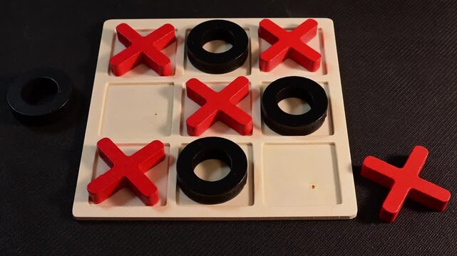 a wooden Tic-Tac-Toe board on a black surface, showing a hand placing a red 'X' piece into the bottom-left corner to win the game.