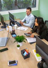 Diverse business professionals sitting at a shared desk, actively collaborating on work tasks and utilizing various technologies in a contemporary office setting with natural light