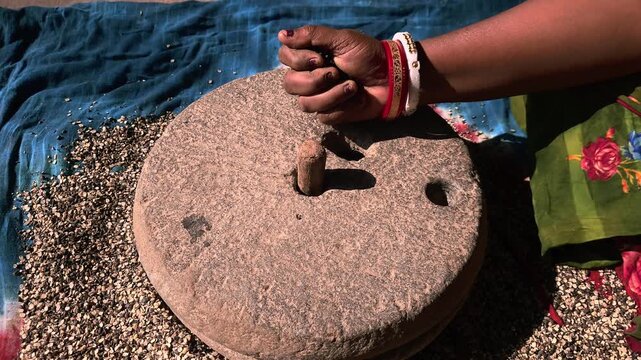 In a rural Indian household, a woman carefully grinds black gram lentils using a traditional hand-operated millstone, preserving age-old food preparation methods passed down through generations.