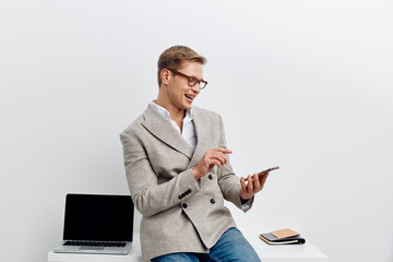 Smiling man using smartphone sitting at modern workplace with laptop and notebooks in bright office environment. Confident professional male engaging with mobile device, casual business attire