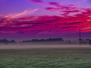 Łąki porośnięte zieloną trawą w letni wieczór. Nad ziemią unosi się warstwa mgły. Niebo jest częściowo pokryte zabarwionymi na czerwono chmurami. © boguslavus
