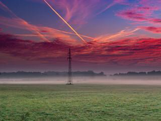 Łąki porośnięte zieloną trawą w letni wieczór. Nad ziemią unosi się warstwa mgły. Niebo jest częściowo pokryte zabarwionymi na czerwono chmurami. © boguslavus