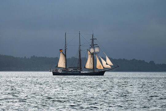 A sailing ship with 3 masts and white sails sailing along the coast in bad weather.