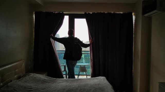 Silhouette in bedroom draws curtains toward city view, remote worker pauses by window before morning routine, soft light filters through fabric, bed in foreground, muted palette evokes calm