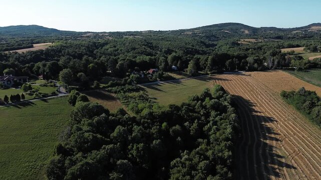 Aerial view of cultivated fields and a small rural village surrounded by forests and rolling hills in summer, showing agricultural patterns, farmland textures and calm countryside atmosphere