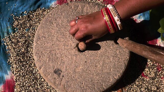 Under natural sunlight in a rural setting, an Indian woman uses a manual grinding stone to process black gram lentils, reflecting cultural traditions and self-reliant domestic practices.