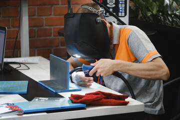 Welder in protective helmet doing manual welding process on two steel plates clamped on work table, demonstrating metal joining technique in workshop environment