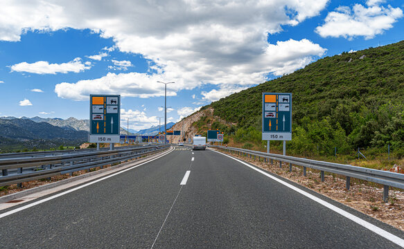 A scenic view of a modern highway in Croatia approaching a toll plaza, flanked by green hills and distant mountains under a blue sky with white clouds.