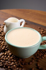 A cup of cappuccino stands on a wooden table among coffee beans, next to it stands a carafe of milk                              