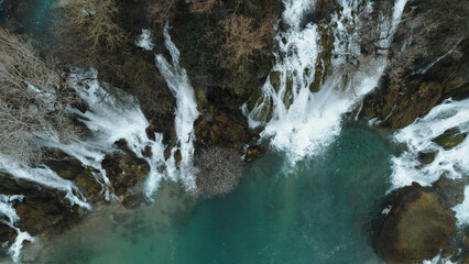 Aerial horizontal view of Kravica Waterfalls in Bosnia during winter, with powerful cascades flowing into a turquoise pool, surrounded by bare trees and rocky cliffs in a serene natural landscape © Ekaterina
