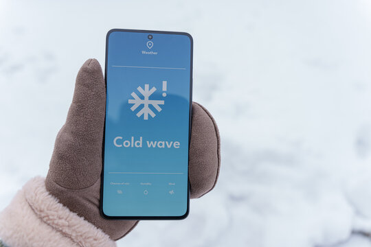 Gloved hand holding smartphone with cold wave alert on screen in snowy winter outdoors