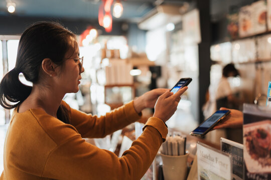 Asian woman customer scanning smartphone to pay at coffee shop counter. Real lifestyle moment for fintech advertising, digital banking apps, QR payment. secure payment messaging, retail technology, - Powered by Adobe
