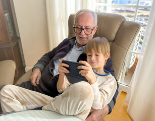 Grandfather enjoys watching his young grandson, looking at the phone together, sitting in the armchair at home.