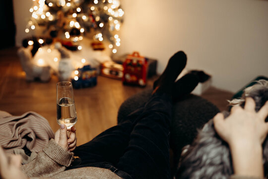 Cozy Christmas evening at home with a woman relaxing on a sofa, holding a small dog, a glass of champagne nearby. Warm festive lights, Christmas tree bokeh, hygge lifestyle and winter holidays comfort - Powered by Adobe