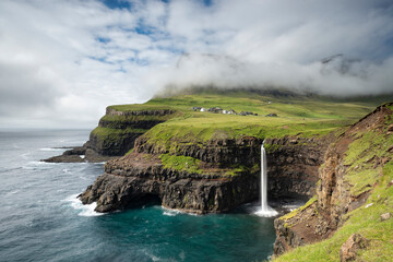 Wide angle shot of Múlafossur waterfall in Gásadalur village, Faroe Islands. Dramatic cliffs meet the Atlantic Ocean under a misty sky. © marcodenaro